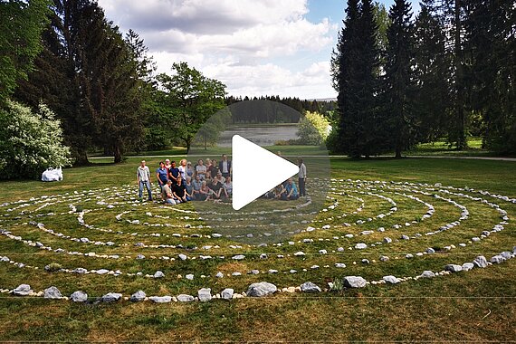 Gruppenfoto von Teilnehmern im Rehazentrum Oberharz, die in einem Steinkreis im Freien stehen. Im Hintergrund sind Bäume und ein See sichtbar.