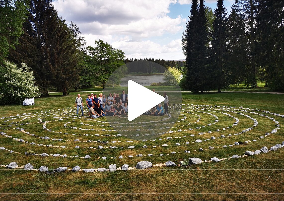 Gruppenfoto von Teilnehmern im Rehazentrum Oberharz, die in einem Steinkreis im Freien stehen. Im Hintergrund sind Bäume und ein See sichtbar.